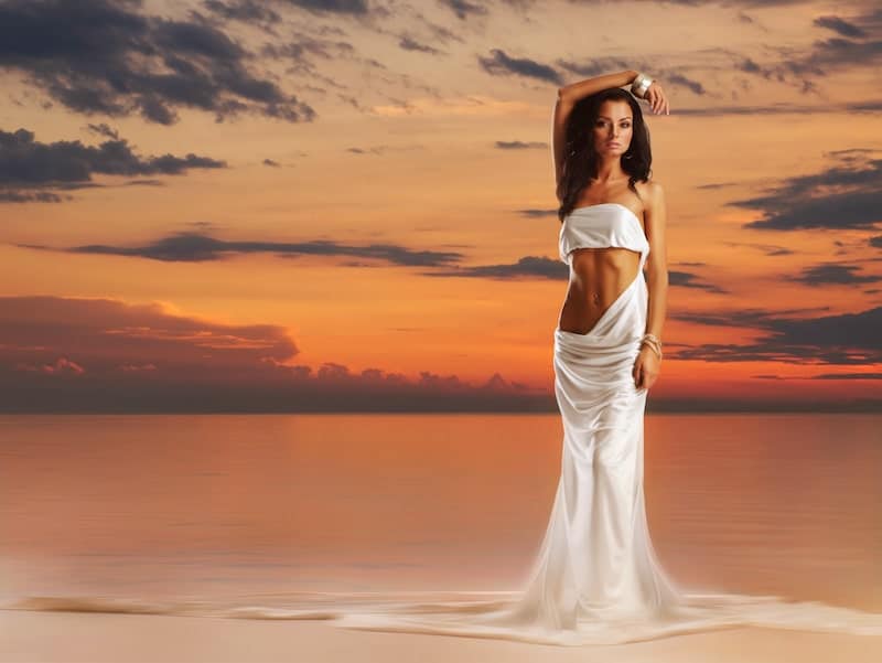 women in white dress standing in front of ocean at sunset
